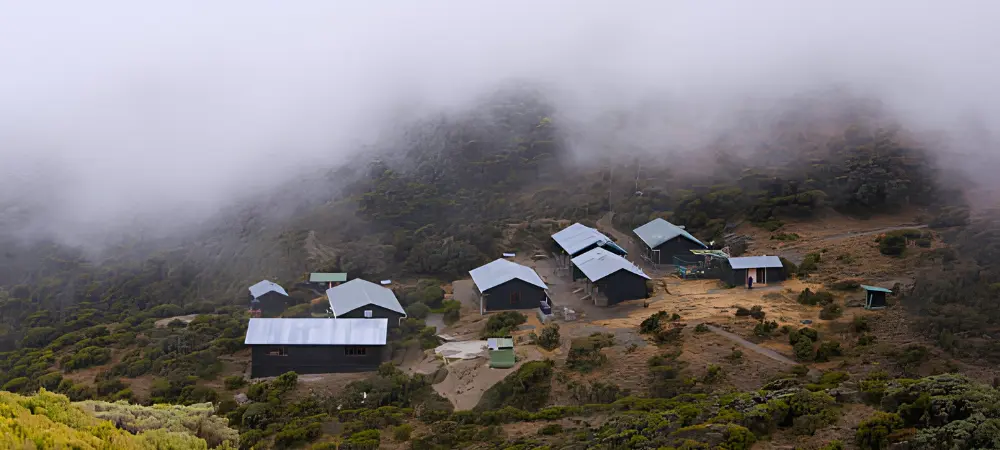 A cluster of mountain huts sits on a rugged hillside of Mount Meru, surrounded by low vegetation and partially covered by thick mist rolling down the slopes.